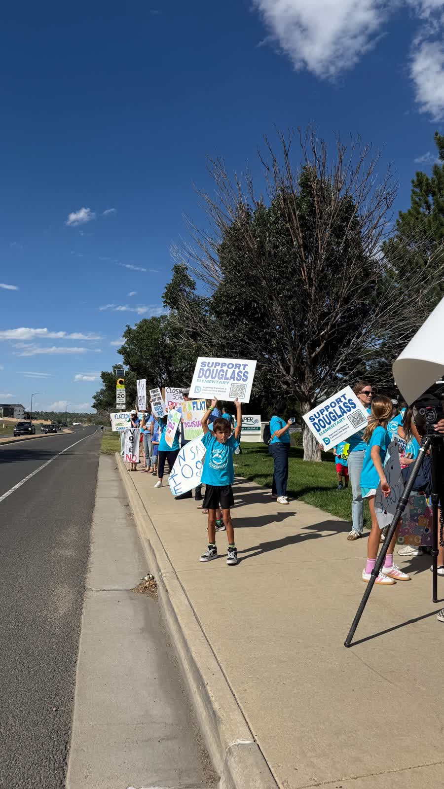 Una fila larga de personas con carteles a la orilla de la calle bajo un cielo azul.